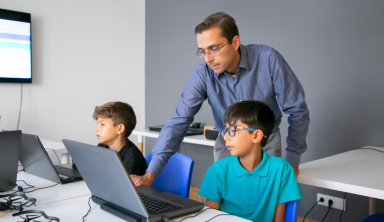 Teacher in glasses standing behind pupil and checking task on laptop